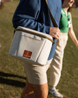 Person holding a white cooler bag with a logo, standing on grass with another person in the background.