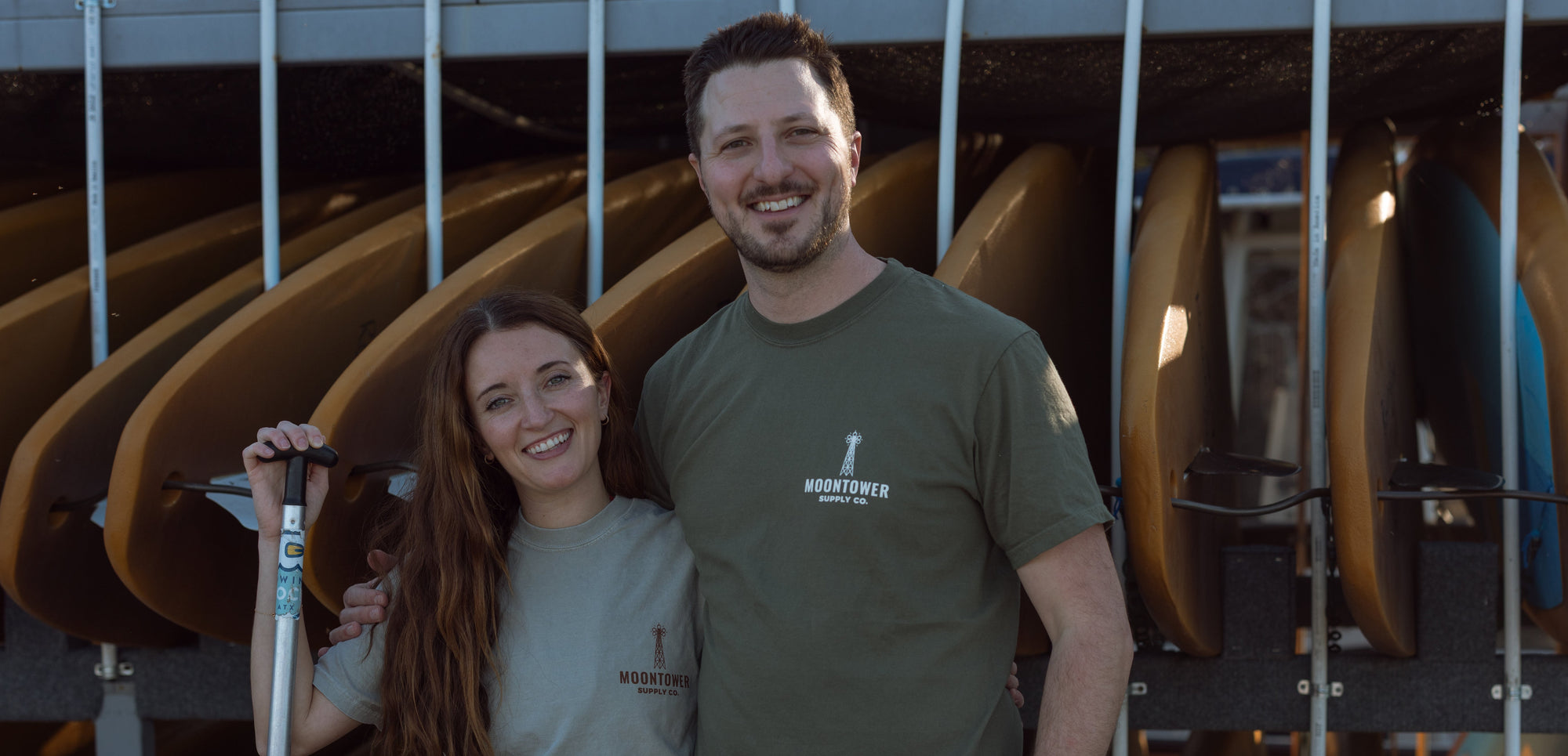 Two people standing in front of kayaks with paddles, wearing matching shirts.